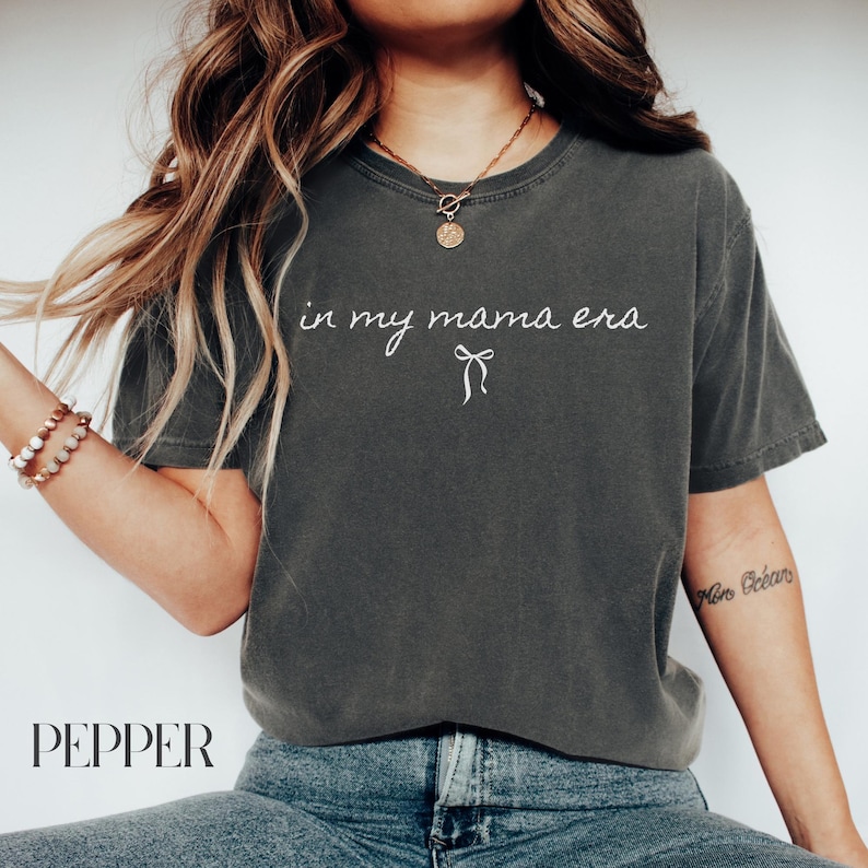May include: A dark gray t-shirt with the text "in my mama era" and a small white bow printed on the front.