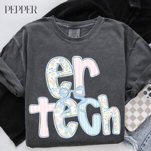 May include: Dark gray long-sleeve shirt with the text "er tech" in a floral and striped design. The letters are outlined in pink and blue, with a light blue bow. The word "PEPPER" is in the top left corner.
