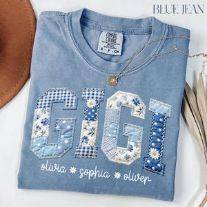 May include: A pale blue Comfort Colors t-shirt featuring the word "GIGI" appliquéd with blue floral and gingham fabrics. The names "olivia", "sophia", and "oliver" are embroidered below. A gold necklace with a pendant lies on the shirt. The text "BLUE JEAN" is at the top.
