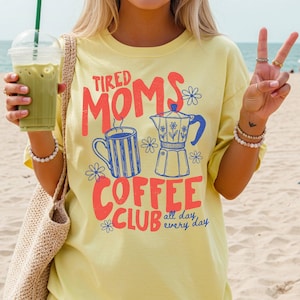 Puede incluir: Camiseta amarilla clara con la frase "Tired Moms Coffee Club" en rojo y naranja, con ilustraciones azules y blancas de una taza de caf&eacute; y una cafetera. La camiseta tiene mangas cortas.