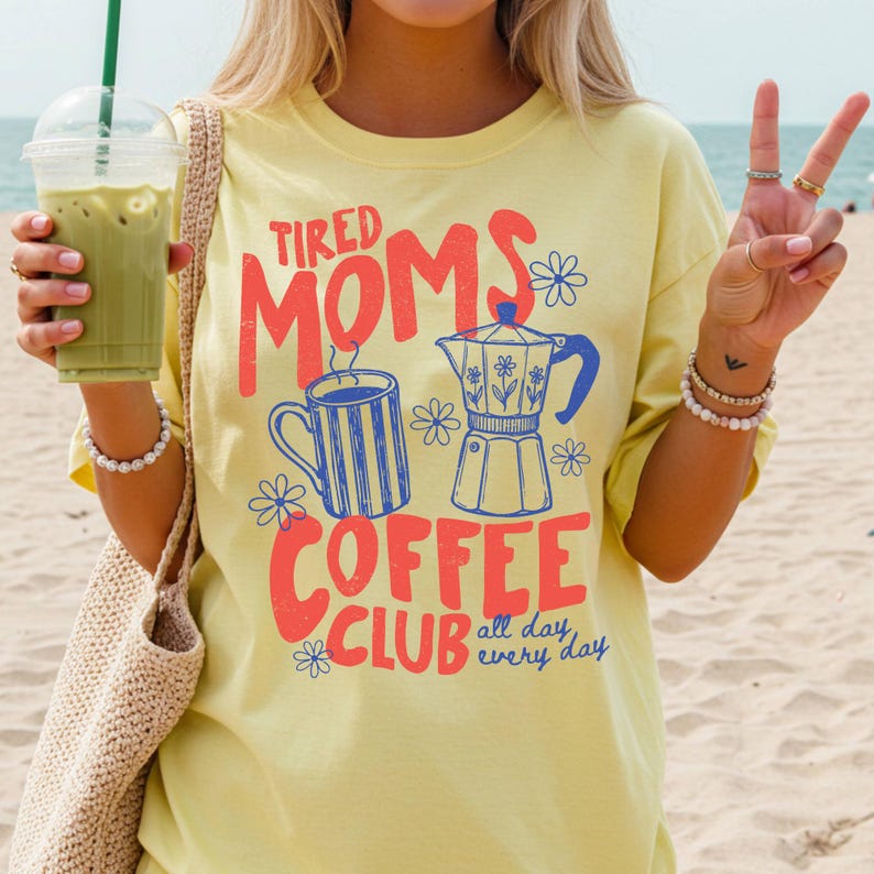 Puede incluir: Camiseta amarilla clara con la frase "Tired Moms Coffee Club" en rojo y naranja, con ilustraciones azules y blancas de una taza de caf&eacute; y una cafetera. La camiseta tiene mangas cortas.