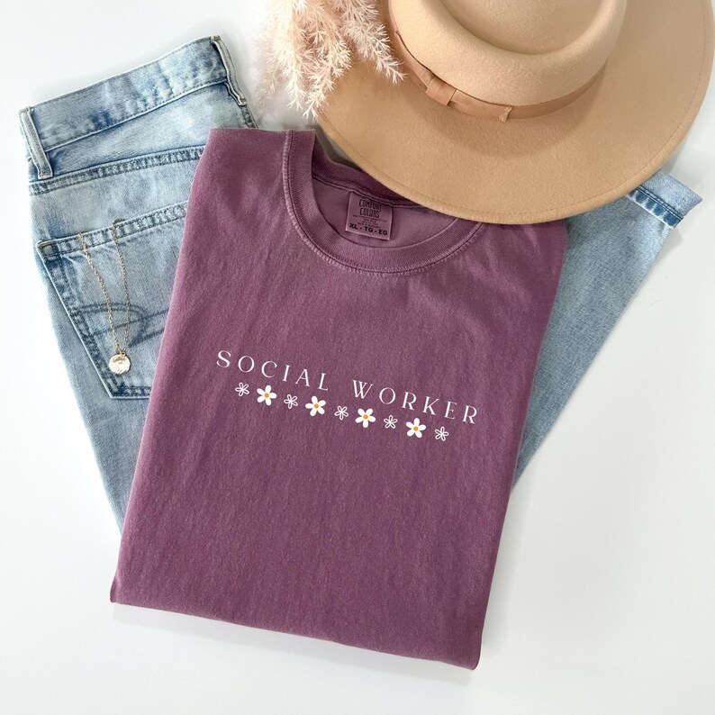 Pu&ograve; includere: Una maglietta color malva con il testo "SOCIAL WORKER" in lettere bianche e piccoli fiori bianchi sotto. La maglietta &egrave; appoggiata su un paio di jeans blu e un cappello marrone.