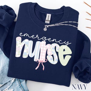 Puede incluir: Sudadera azul marino con la frase "emergency nurse". La palabra "nurse" est&aacute; decorada con estampados florales, cuadros y rayas. Un collar plateado con un colgante est&aacute; sobre la sudadera.
