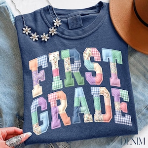 Peut inclure: Un sweat-shirt bleu marine avec l'inscription "FIRST GRADE" en lettres patchwork colorées. Un collier floral et un chapeau marron sont visibles. Le sweat-shirt est en matière douce, parfait pour la rentrée.