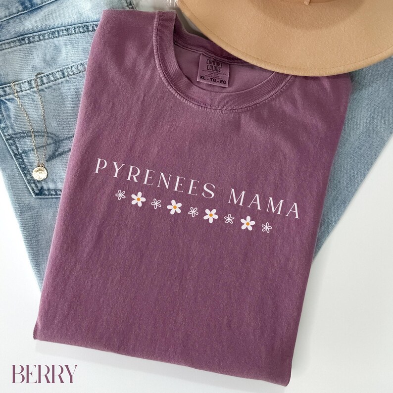 Puede incluir: Una camiseta de color malva con el texto "PYRENEES MAMA" en letras blancas y una fila de margaritas blancas debajo del texto.