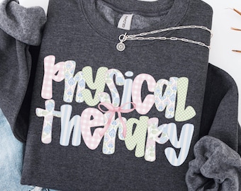 May include: Dark heather gray sweatshirt with the words "Physical Therapy" in pastel colors and patterns. A silver necklace with a sun pendant is draped across the shirt. The text "DARK HEATHER" is at the bottom.