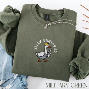 May include: Military green sweatshirt with the text "SILLY ENGINEER" surrounding an embroidered cartoon goose wearing a yellow hard hat and holding tools. A silver necklace with a small pendant is on the sweatshirt. The words "MILITARY GREEN" are at the bottom.