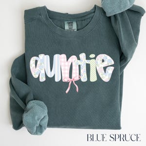 May include: Blue spruce sweatshirt with the word "auntie" in a whimsical font. The letters are decorated with floral and gingham patterns, and a pink bow is centered below the word. The sweatshirt is a Comfort Colors brand.