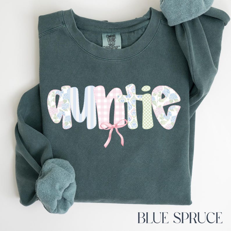 May include: Blue spruce sweatshirt with the word "auntie" in a whimsical font. The letters are decorated with floral and gingham patterns, and a pink bow is centered below the word. The sweatshirt is a Comfort Colors brand.