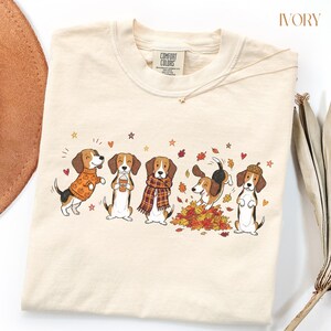 Puede incluir: Una camiseta de color marfil con un gr&aacute;fico de cinco perros beagle con ropa y actividades de oto&ntilde;o, incluyendo un su&eacute;ter de calabazas, una taza de caf&eacute;, una bufanda de cuadros, jugando entre hojas secas y con un sombrero de bellota.