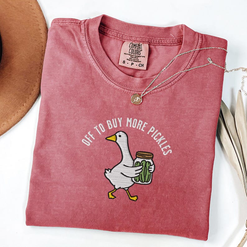 May include: A dusty rose-colored t-shirt with the phrase "OFF TO BUY MORE PICKLES" embroidered above a cartoon goose holding a jar of pickles. A silver necklace with a pendant is draped across the shirt.