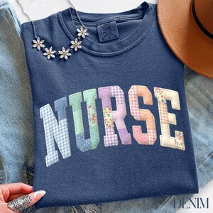 Peut inclure: Sweat-shirt bleu marine avec le mot "NURSE" en lettres patchwork. Les lettres sont dans différentes couleurs pastel et motifs. Un collier floral argenté et un chapeau marron sont également visibles.