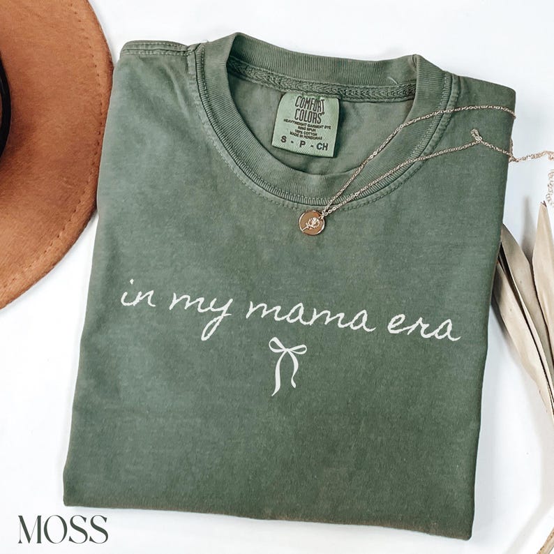 May include: A green t-shirt with the text "in my mama era" and a small bow underneath. The shirt is folded and has a white tag with the text "COMFORT COLORS" on it.