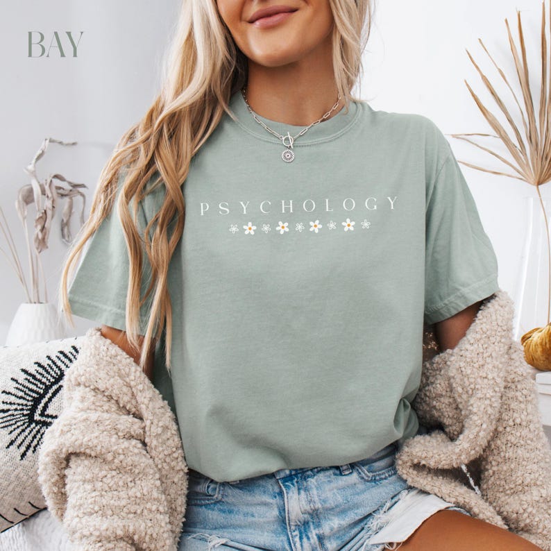 May include: A sage green t-shirt with the text "Psychology" in white lettering and a row of small white flowers underneath. The shirt is being worn by a person with long blonde hair.