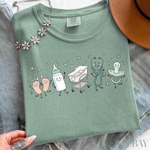 May include: A sage green t-shirt featuring a graphic print of cartoon baby items including feet, a bottle, a bassinet, a stethoscope, and a pacifier, all with smiling faces. The shirt has a crew neckline and a Comfort Colors brand label inside.