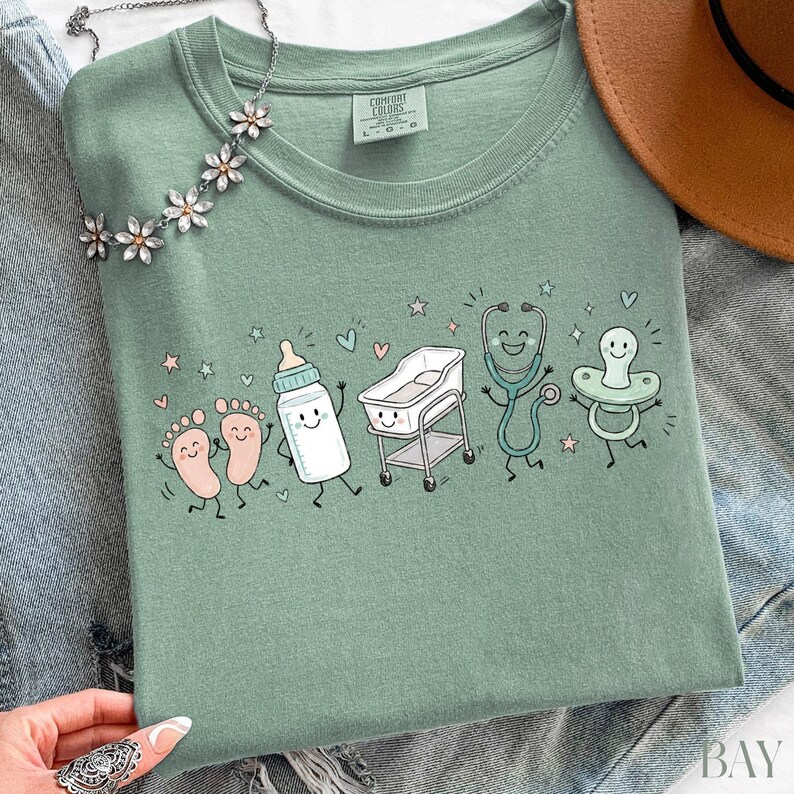 May include: A sage green t-shirt featuring a graphic print of cartoon baby items including feet, a bottle, a bassinet, a stethoscope, and a pacifier, all with smiling faces. The shirt has a crew neckline and a Comfort Colors brand label inside.