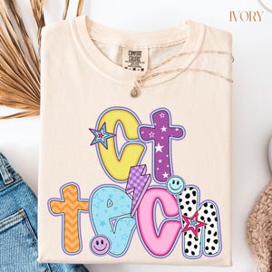 May include: Ivory-colored sweatshirt with the words "Get Teach" in colorful, playful lettering. The design includes stars, a lightning bolt, and smiley faces. The sweatshirt is folded and has a Comfort Colors tag.