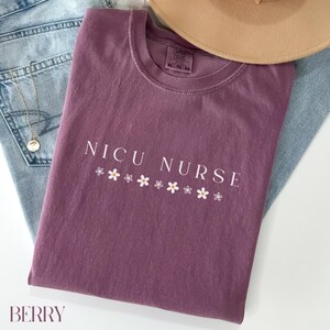 Peut inclure: Un t-shirt mauve avec le texte "NICU NURSE" en lettres blanches et une rang&eacute;e de marguerites blanches sous le texte.