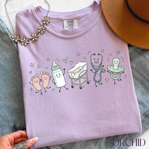 May include: A light purple t-shirt featuring a graphic print of cartoon baby items including feet, a bottle, a bassinet, a stethoscope, and a pacifier, all with smiling faces. The shirt has a Comfort Colors label at the inner neckline.