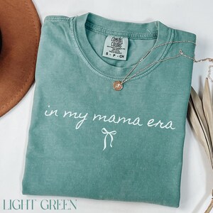 May include: A light green t-shirt with the text "in my mama era" in white lettering with a white bow underneath. The shirt is made of a soft, comfortable material.