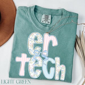 May include: Light green t-shirt with the word "ER TECH" in a floral and striped design. The letters are decorated with a floral pattern, pink stripes, and blue stripes. A light blue bow is between the letters "ER". The shirt has a Comfort Colors tag.