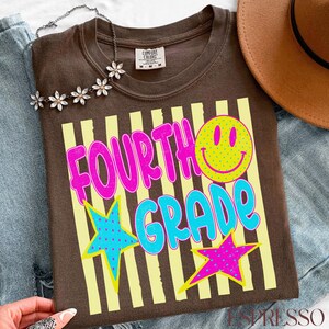 Op de afbeelding: Een bruin T-shirt met de woorden "FOURTH GRADE" in roze en blauw, een gele smiley en twee sterren. Het shirt heeft gele verticale strepen en een Comfort Colors label.