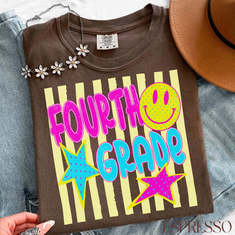 Op de afbeelding: Een bruin T-shirt met de woorden "FOURTH GRADE" in roze en blauw, een gele smiley en twee sterren. Het shirt heeft gele verticale strepen en een Comfort Colors label.