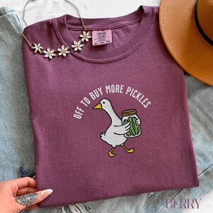 May include: A maroon Comfort Colors t-shirt with the text "OFF TO BUY MORE PICKLES" encircling a cartoon goose carrying a jar of pickles. The goose is white with yellow feet and a yellow beak. A floral necklace and a brown hat are also visible.