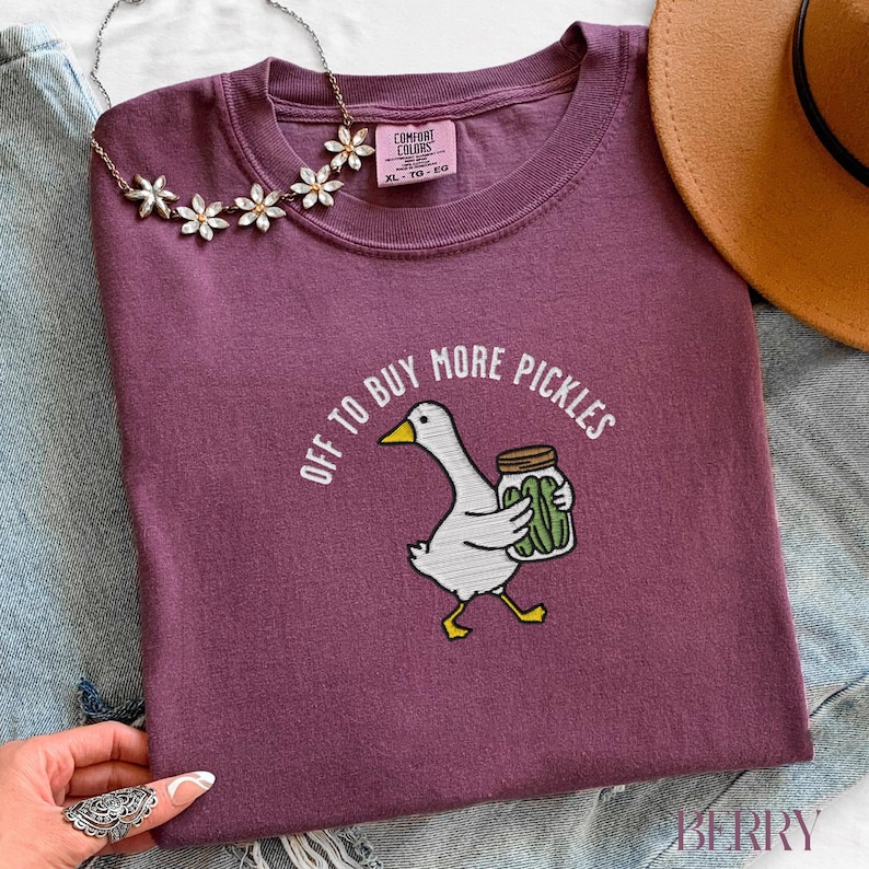 May include: A maroon Comfort Colors t-shirt with the text "OFF TO BUY MORE PICKLES" encircling a cartoon goose carrying a jar of pickles. The goose is white with yellow feet and a yellow beak. A floral necklace and a brown hat are also visible.