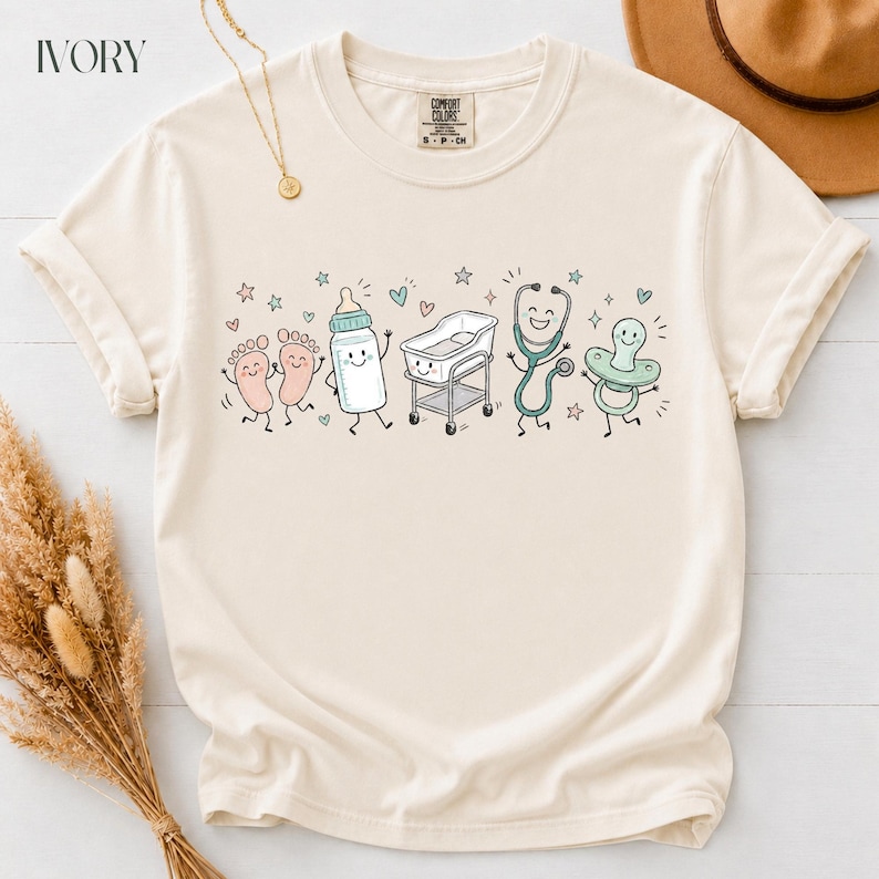 May include: An ivory short-sleeve t-shirt featuring a graphic print of cartoon baby items including feet, a bottle, a bassinet, a stethoscope, and a pacifier. The design includes small stars and hearts in muted pastel colors across the chest area.