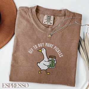 May include: Espresso-colored t-shirt with the phrase "OFF TO BUY MORE PICKLES" and a cartoon goose carrying a jar of pickles. The shirt has a crew neck and short sleeves. A silver necklace with a pendant is draped across the shirt.
