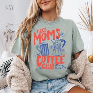 Puede incluir: Camiseta verde salvia con el texto "Tired Moms Coffee Club" en rojo y azul. El dise&ntilde;o incluye una taza de caf&eacute;, una cafetera y detalles florales. La camiseta es de corte holgado.