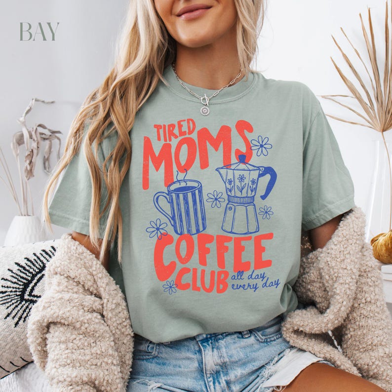 Puede incluir: Camiseta verde salvia con el texto "Tired Moms Coffee Club" en rojo y azul. El dise&ntilde;o incluye una taza de caf&eacute;, una cafetera y detalles florales. La camiseta es de corte holgado.