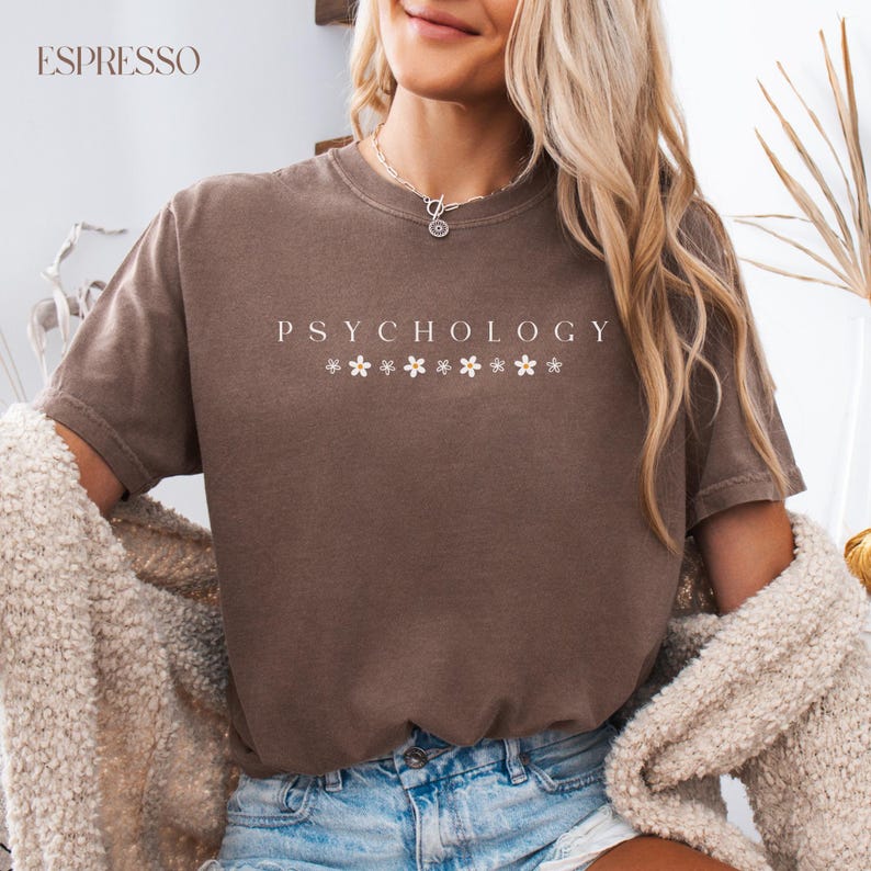 May include: A brown t-shirt with the text "Psychology" printed in white with small white flower accents. The model is wearing blue jeans.