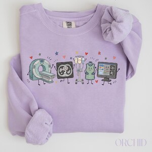 May include: A light purple crewneck sweatshirt featuring a graphic print of cartoon medical equipment including a CT scanner, an X-ray, an IV pole, a lead apron, and a computer monitor, all with smiling faces, surrounded by small colorful hearts and stars.
