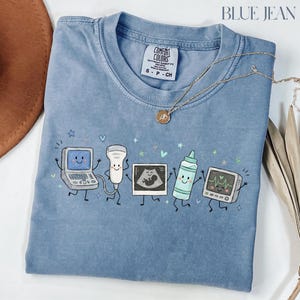 May include: A blue denim-colored t-shirt featuring a graphic print of five anthropomorphic medical devices including an ultrasound machine, a probe, a sonogram image, a gel bottle, and a heart monitor, all with smiling faces and thin limbs.