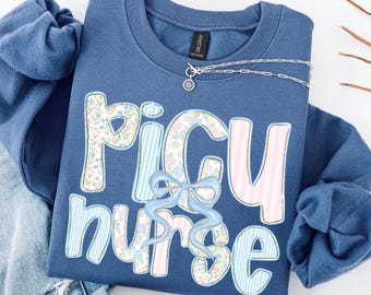 May include: Indigo blue sweatshirt with the words "PICU nurse" in a decorative font. The letters are decorated with floral patterns, pink stripes, and blue stripes. A light blue bow is centered below the letters. A silver necklace with a small pendant is draped across the sweatshirt.