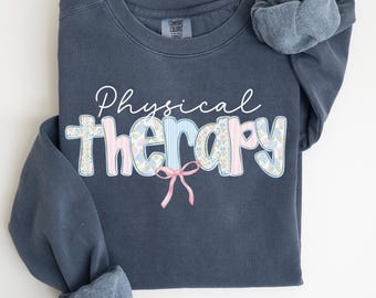 May include: A denim blue sweatshirt with the words "Physical Therapy" in white script and floral and striped lettering. A pink bow is below the word "therapy". The sweatshirt has a soft, fleece-lined interior and the word "DENIM" is printed in the bottom right corner.