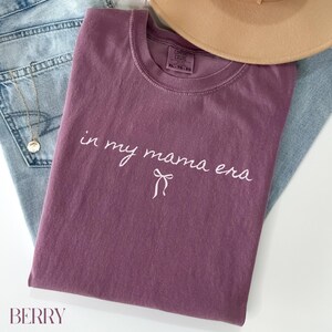 May include: A mauve colored t-shirt with the text "in my mama era" in white cursive lettering with a small white bow underneath.