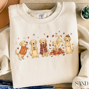 May include: A cream-colored crewneck sweatshirt featuring a graphic print of five golden retriever dogs in various autumn-themed poses. The dogs are depicted wearing sweaters, scarves, and an acorn hat, surrounded by colorful fall leaves and small hearts.