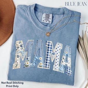 May include: A pale blue t-shirt with the word "MAMA" in patchwork letters. The letters feature floral, gingham, and polka dot patterns in shades of blue and white. A gold necklace with a pendant rests on the shirt. The text "BLUE JEAN" is at the top.