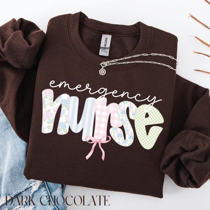 Puede incluir: Una sudadera de color chocolate oscuro con las palabras "emergency nurse" en escritura blanca. La palabra "nurse" tiene un dise&ntilde;o de patchwork con estampados florales, a rayas y de cuadros. Un collar plateado con un colgante est&aacute; sobre la sudadera.