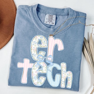 May include: A blue Comfort Colors t-shirt with the text "ER TECH" in a floral and striped design. The shirt is folded, with a small tag at the top. A gold necklace is draped across the shirt.