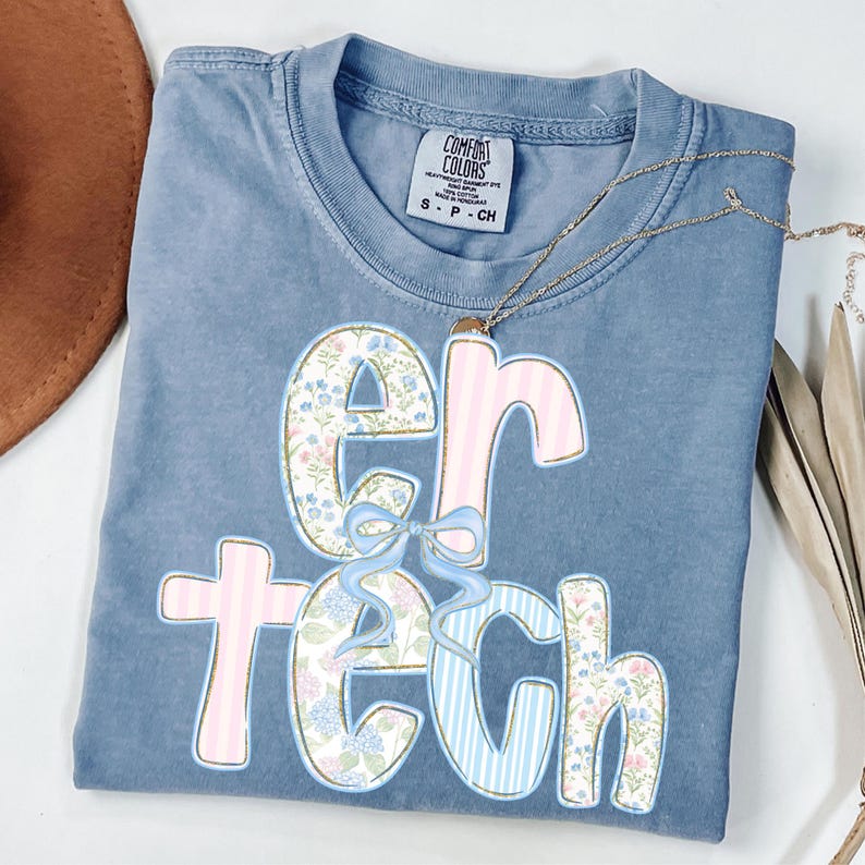 May include: A blue Comfort Colors t-shirt with the word "ER TECH" in a floral and striped design. The shirt has a small tag at the top and is folded neatly. A gold necklace is draped across the shirt.