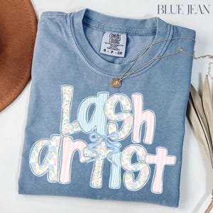 May include: Blue jean colored t-shirt with the words "Lash Artist" in a floral and striped design. The shirt has a crew neck and a small tag at the top. A gold necklace with a pendant is draped across the shirt.