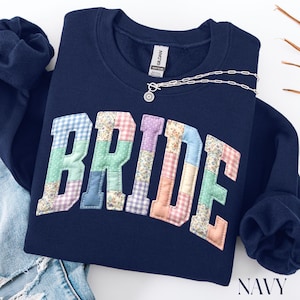 May include: Navy blue sweatshirt with the word "BRIDE" appliqued across the front in a patchwork design. The patchwork letters feature various pastel floral and gingham patterns. A silver necklace with a sunburst pendant rests on the sweatshirt.