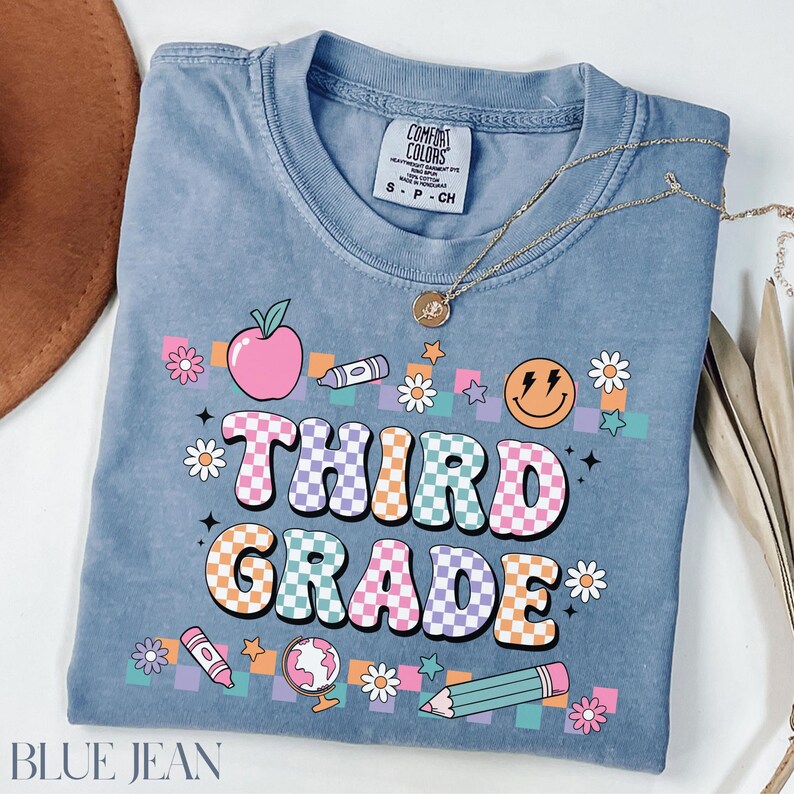 May include: A light blue t-shirt with a checkered design that says "Third Grade" in colorful letters. The shirt is decorated with a variety of school-themed graphics, including an apple, a crayon, a pencil, a globe, and a smiley face.