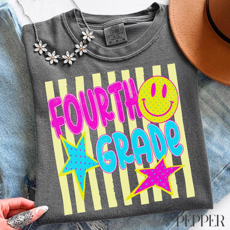 Op de afbeelding: Donkergrijs T-shirt met de woorden "FOURTH GRADE" in roze en blauw, een gele smiley en twee sterren. Het shirt heeft een gele gestreepte achtergrond. Een zilveren ketting en een bruine hoed zijn zichtbaar.