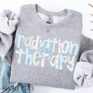 May include: Sport grey sweatshirt with the words "Radiation Therapy" in a floral and striped design. The text is in light blue, pink, and floral patterns, with a light blue bow. A silver necklace is on the sweatshirt.