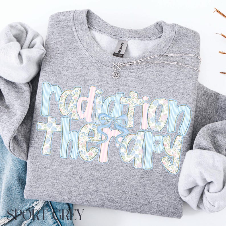 May include: Sport grey sweatshirt with the words "Radiation Therapy" in a floral and striped design. The text is in light blue, pink, and floral patterns, with a light blue bow. A silver necklace is on the sweatshirt.